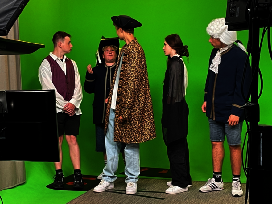 Fünf Jugendliche stehen in historischen Kostümen (Dreispitz, Perücke, Mäntel) vor einem Greenscreen in einem Filmstudio bei Dreharbeiten.