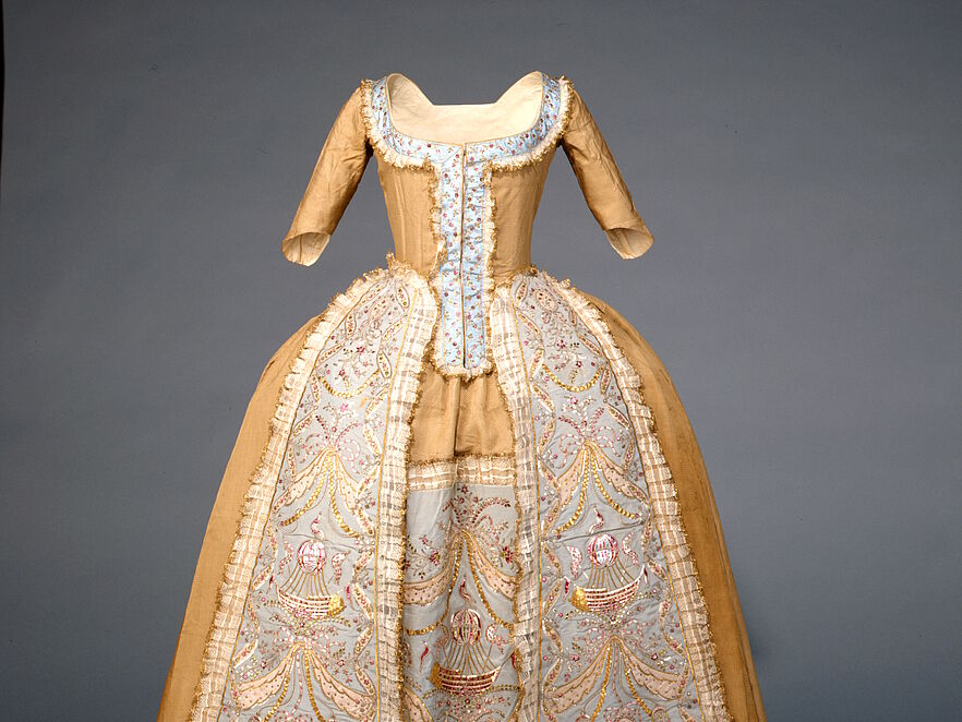 Frontansicht einer goldbeigen Robe à la française aus dem 18. Jahrhundert vor grauem Hintergrund. Kleid ist als historisches Ausstellungsobjekt ohne Trägerin präsentiert. Hellblauer Unterrock mit Stickereien von pinkfarbenen Heißluftballons, Schleifen und Goldgirlanden, eng tailliertes Mieder, weit ausgestellter Rock über Reifrock-Konstruktion.