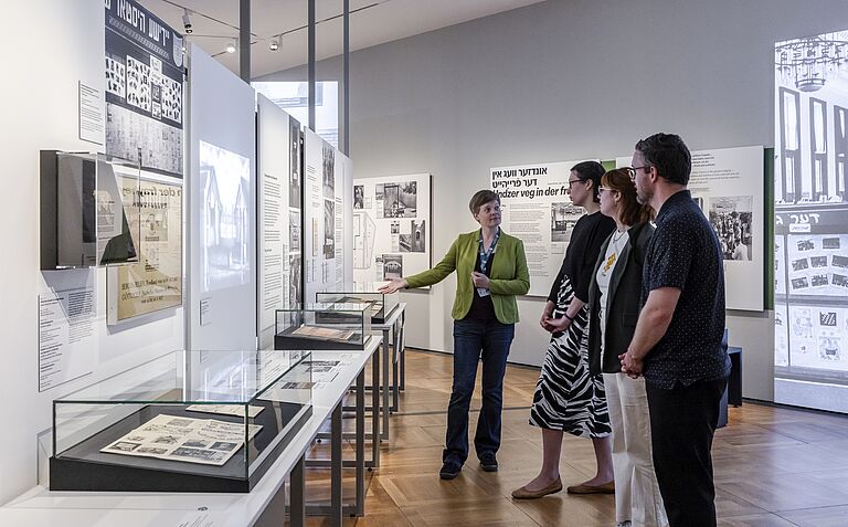 Ausstellungsansicht „Gewalt ausstellen: Erste Ausstellungen zur NS-Besatzung in Europa, 1945-1948“ Situation einer Führung in Ausstellung „Gewalt ausstellen: Erste Ausstellungen zur NS-Besatzung in Europa, 1945-1948“ mit einer weiblichen Person als Guide und drei Teilnehmenden