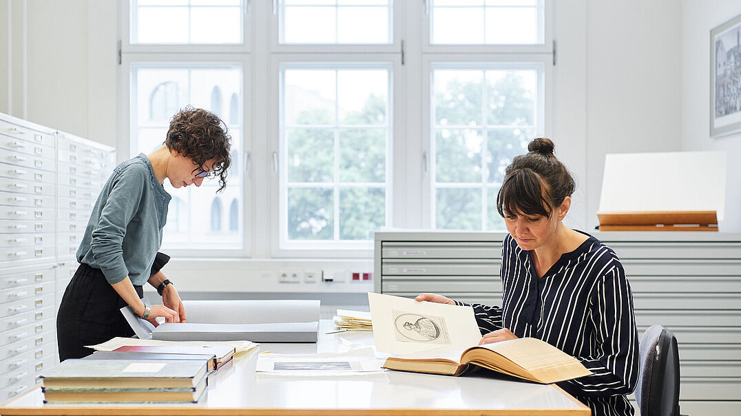 Zwei Personen arbeiten an einem Tisch in einem gut beleuchteten Raum mit großen Fenstern. Einer prüft Dokumente, während der andere ein Buch liest.