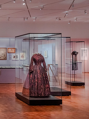 Blick in die Ausstellung "Richard Wagner und das deutsche Gefühl". Neben verschiedenen Grafiken in Rahmen ist vor allem ein dunkles geblümtes Kleid präsent in einer Vitrine zu sehen.