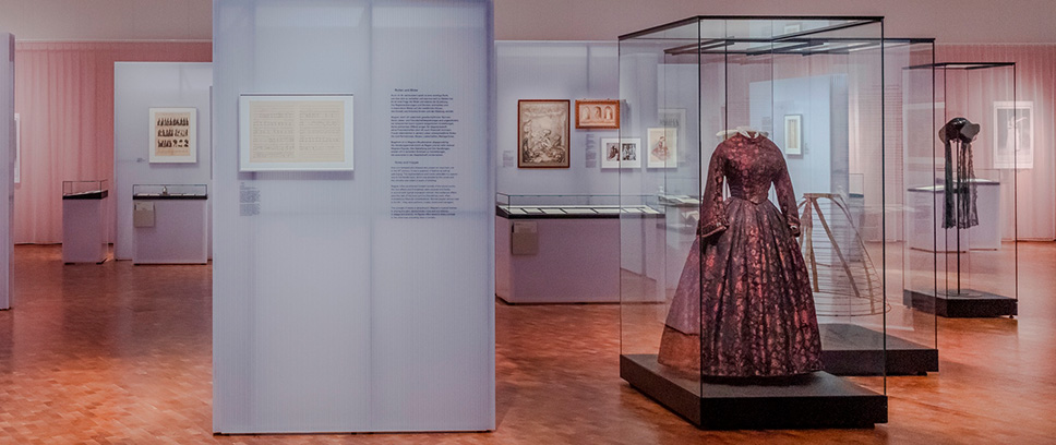 Blick in die Ausstellung "Richard Wagner und das deutsche Gefühl". Neben verschiedenen Grafiken in Rahmen ist vor allem ein dunkles geblümtes Kleid präsent in einer Vitrine zu sehen.