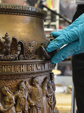 An der Bronzesäule sind Figuren, Adler, und Brustpanzer zu sehen. Außerdem ein Spruchband was drumherum läuft. Eine mit einem blauen Handschuh bekleidete Hand hält einen der Adlerappliken fest.