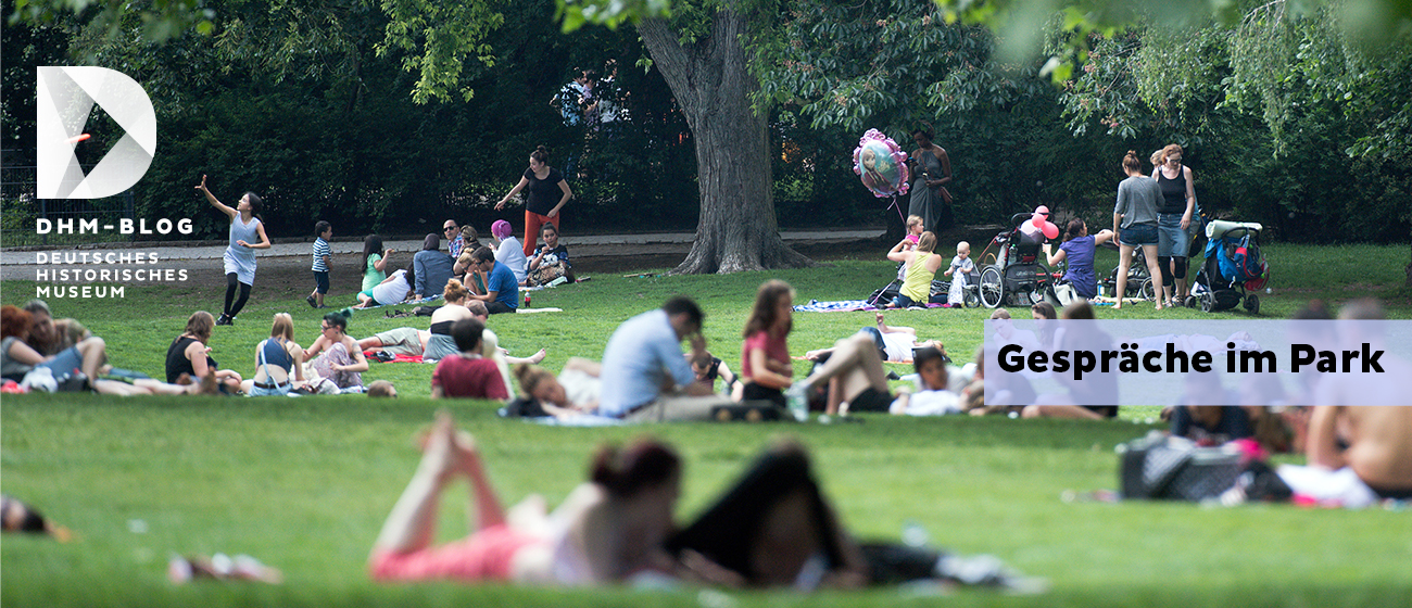 Auf dem Foto ist ein Park zu sehen. In dem park befinden sich viele Menschen, die sitzen, stehen oder rumlaufen. Auf dem Bild steht "Gespräche im Park auf der linken Seite"