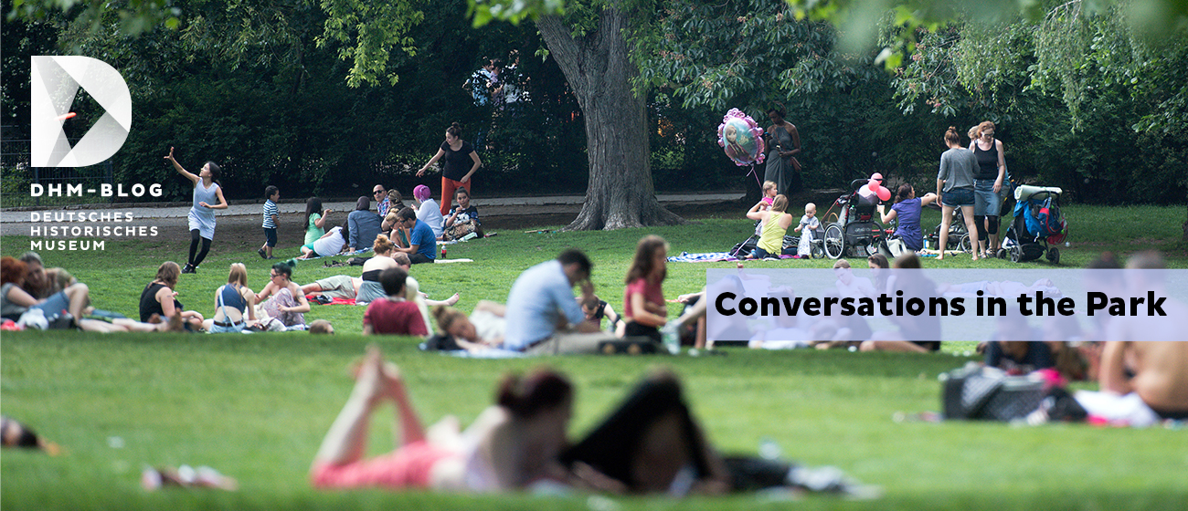 Auf dem Foto ist ein Park zu sehen. In dem park befinden sich viele Menschen, die sitzen, stehen oder rumlaufen. Im textfeld steht "Conversations in the park"