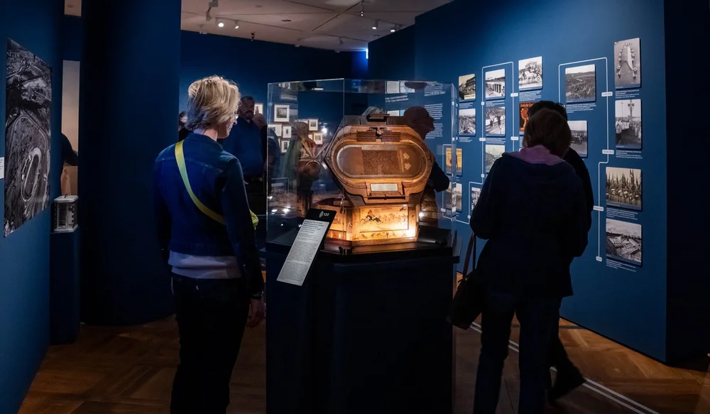 Ausstellungsansicht der Ausstellung „Objekte. Geschichte. Geschichten. Blick in die Sammlung“. In der Mitte des Bildes ist eine Vitrine zu sehen, darin steht ausgeleuchtet eine hexagonale Truhe mit einem Modell des Deutschen Stadions in Berlin-Charlottenburg von Gustav Baumann (von 1912 bzw. 1923/24). Im Hintergrund stehen einige Personen, die einen Zeitstrahl erkunden.