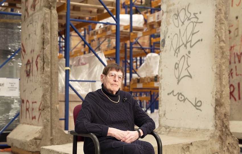 Eine Frau im Rollstuhl sitzt in einer Halle vor Teilen der Berliner Mauer