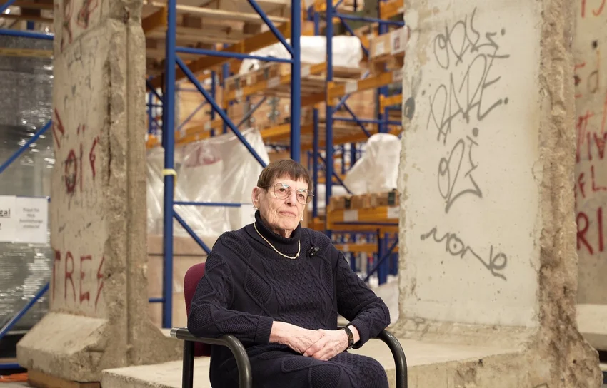 [Translate to English:] Eine Frau im Rollstuhl sitzt in einer Halle vor Teilen der Berliner Mauer