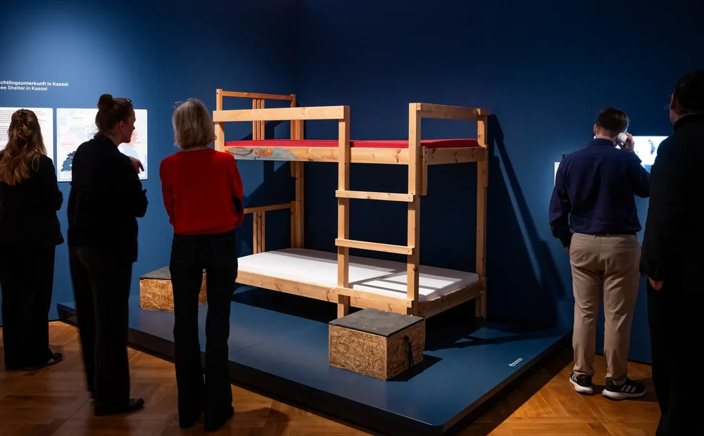 Ausstellungsgäste betrachten ein Doppelstockbett, das in einer Unterkunft für Geflüchtete stand. Es handelt sich um Holzbetten des Herstellers Ikea.