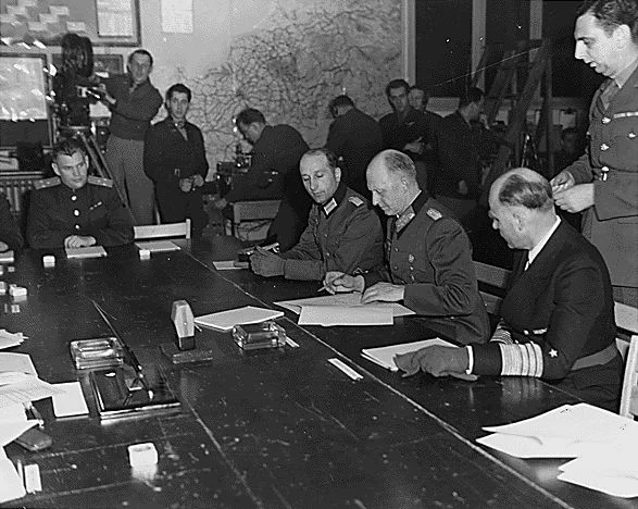 Black-and-white photograph: Colonel-General Alfred Jodl is sitting at a table. Surrounded by several people, he is signing a document lying in front of him.