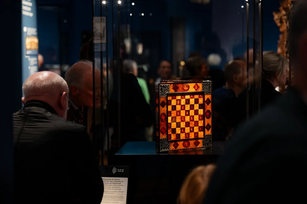 Das Bild zeigt eine relativ dunkle Ausstellungsansicht von „Objekte. Geschichte. Geschichten. Blick in die Sammlung“ mit dem Fokus auf eine Brettspielkassette für Mühle, Schach und Tric-Trac, die u.a. aus Bernstein, Ebenholz und Silber gefertigt ist.
