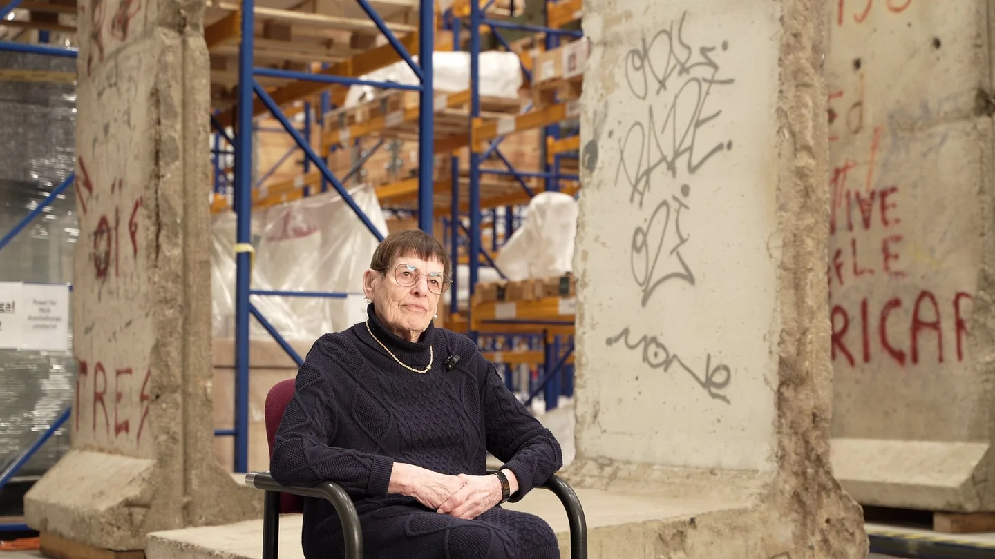 Eine Frau im Rollstuhl sitzt in einer Halle vor Teilen der Berliner Mauer