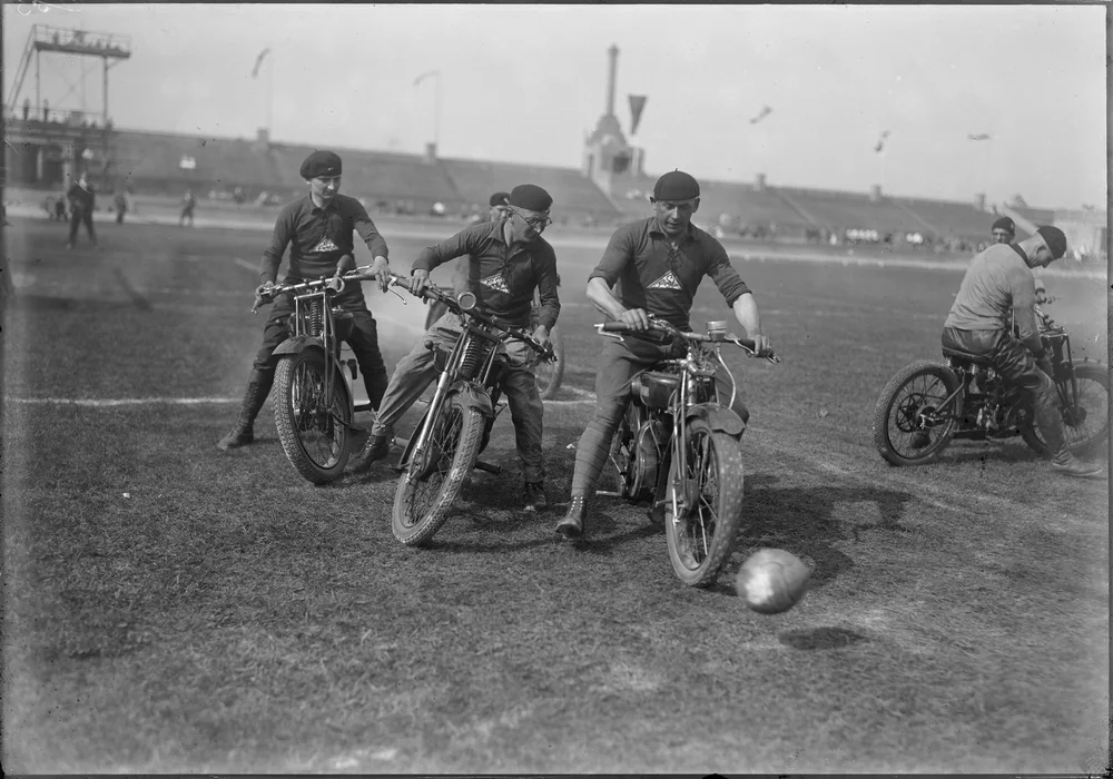 Schwarz-Weiß-Fotografie: Drei Motorradfahrer fahren auf einem Rasenplatz einem Ball hinterher. Im Hintergrund sind Tribünen und Zuschauer zu sehen.