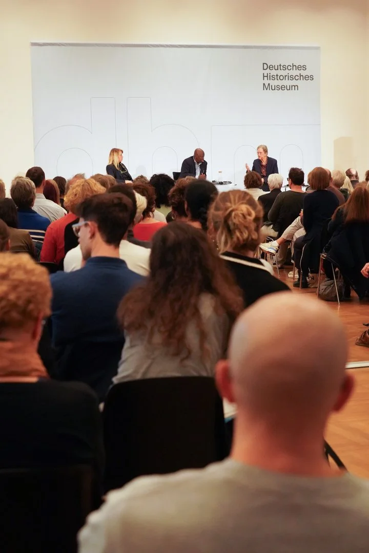 Raumansicht der Veranstaltung: Im Vordergrund ist das Publikum von hinten zu sehen, im Hintergrund die Bühne mit den Podiumsgästen