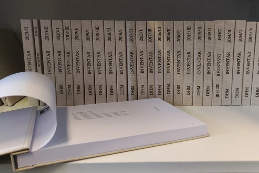 [Translate to English:] Das Bild zeigt einige in Leinen gebundene Inventarbücher des DHM, die aufgereiht nebeneinander stehen, sortiert nach Jahren von 2013 bis 2021. Eines der Inventarbücher liegt aufgeschlagen im Vordergrund und zeigt das Inhaltsverzeichnis.