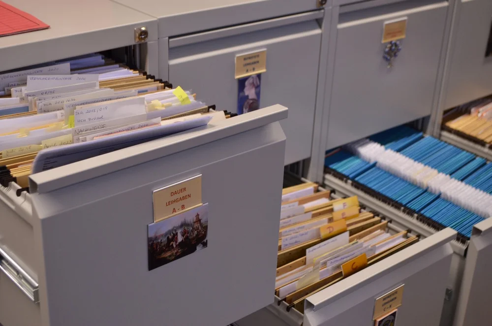 Ein hellgrauer Aktenschrank mit mehreren Schubläden, wovon eine geöffnet ist und den Blick freigibt auf zahlreiche, geordnete Akten und Hängeregistern.