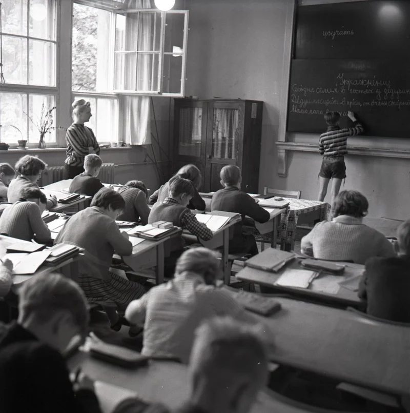 Schwarz-weiß Fotografie mit Kindern in einem Klassenzimmer, Blick auf die Tafel auf der die Lehrerin schreibt