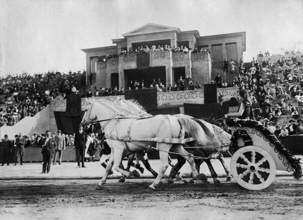 Schwarz-Weiß-Fotografie: Ein festlich geschmückter Pferdewagen mit mehreren weißen Pferden fährt an dicht besetzten Tribünen vorbei.