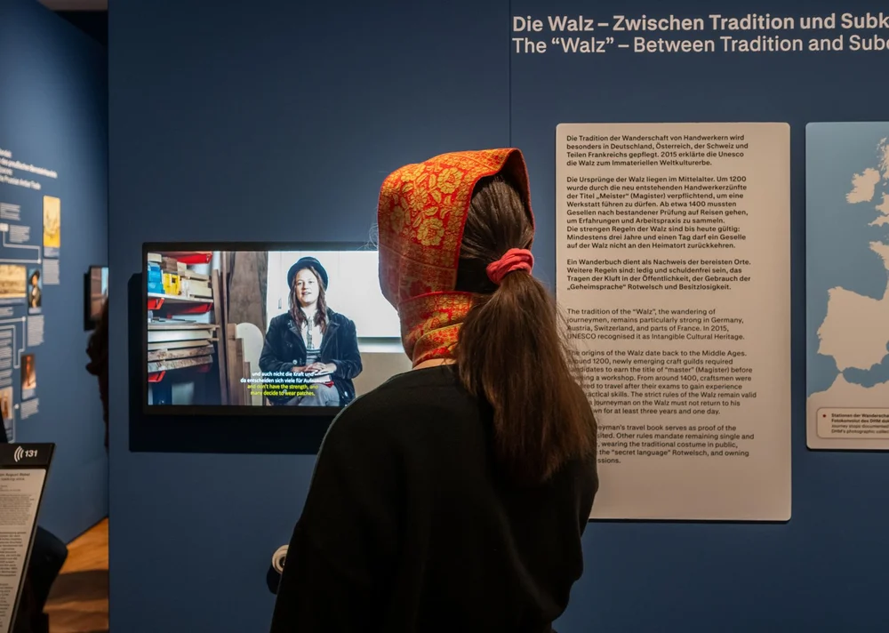 Eine Besucherin steht in der Ausstellung und schaut sich ein Videointerview einer Wandergesellin an.