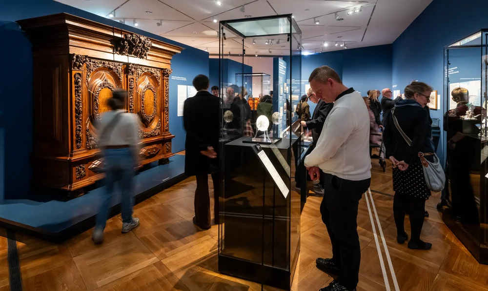 Ausstellungsansicht von „Objekte. Geschichte. Geschichten. Blick in die Sammlung“, auf der mehrere Personen zu sehen sind, die durch die Ausstellung laufen. Ein Herr im Vordergrund betrachtet ein Objekt in einer Vitrine: ein graviertes Straußenei mit Motiven aus Jan Huygen van Linschotens Itinerario (Niederlande, ca. 1600/1620)