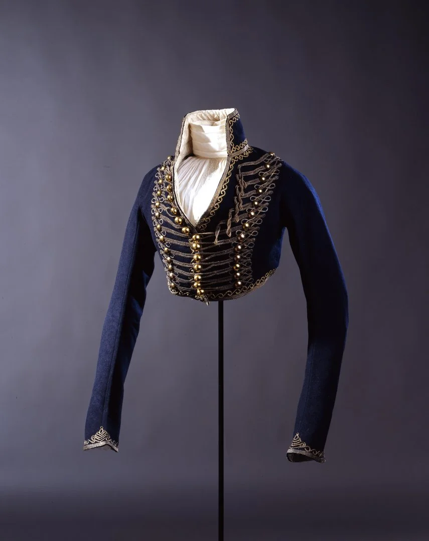 Das Foto zeigt eine Installationsansicht einer preußischblauen, kurzen Reitjacke mit Stehkragen und weißer Fütterung. Brustbereich und Säume sind mit Goldknöpfen und -stickereien verziert.