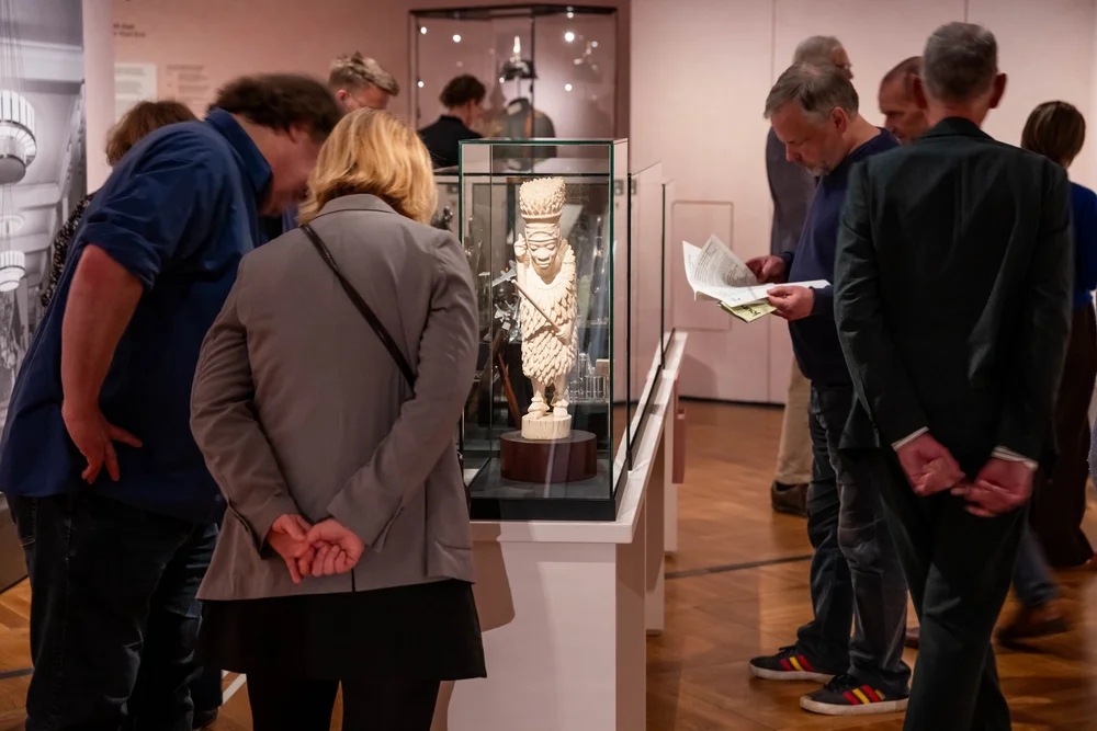 Auf dem Foto sieht man einige Gäste, die in der Ausstellung „Objekte. Geschichte. Geschichten. Blick in die Sammlung“ gebeugt über Vitrinen stehen oder durch die Ausstellung laufen.