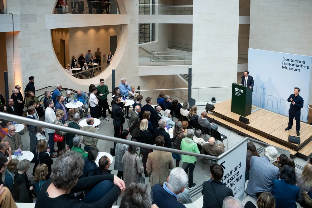 Impressionen der Ausstellungseröffnung „Objekte. Geschichte. Geschichten. Blick in die Sammlung“: DHM-Präsident Raphael Gross steht auf der Bühne und begrüßt die Gäste, die im Foyer auf allen Etagen verteilt stehen und auf die Bühne blicken.