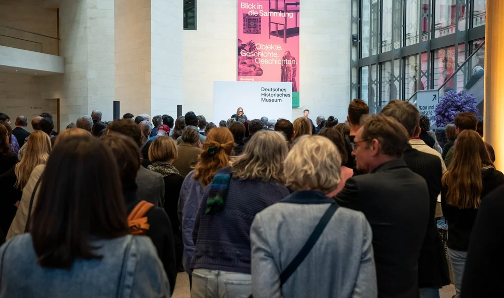 Blick aus dem Foyer im Erdgeschoss auf die Bühne über die Köpfe der stehenden Gäste hinweg. In der Ferne ist auf der Bühne am Rednerpult die Gastrednerin Charlotte Klonk zu erkennen.
