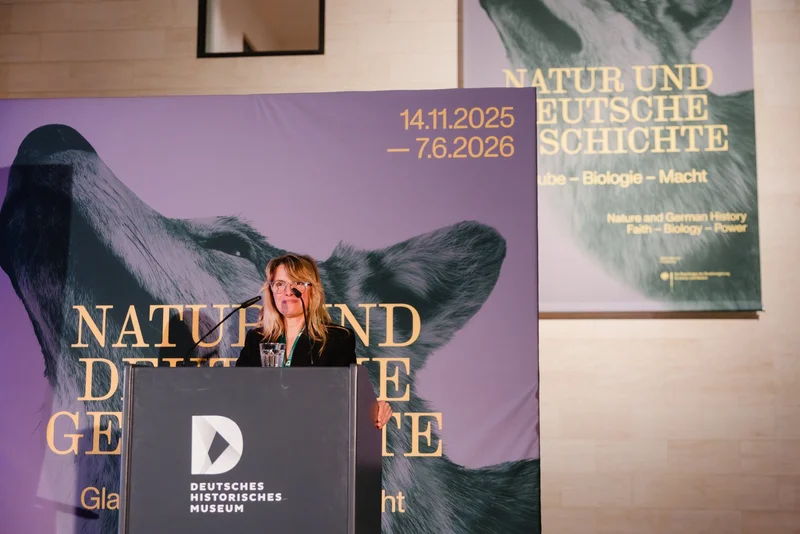 [Translate to English:] Kuratorin Julia Voss spricht auf einem Podium. Sie trägt einen schwarzen Blazer und hat lange blonde Haare. Im Hintergrund ist das Plakatmotiv zur Ausstellung mit einem Wolf zu sehen. 