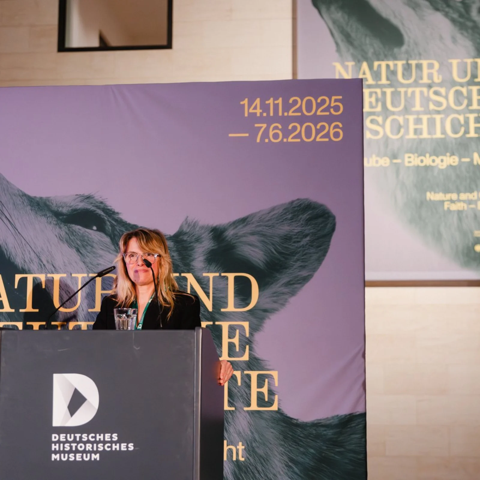 [Translate to English:] Kuratorin Julia Voss spricht auf einem Podium. Sie trägt einen schwarzen Blazer und hat lange blonde Haare. Im Hintergrund ist das Plakatmotiv zur Ausstellung mit einem Wolf zu sehen. 