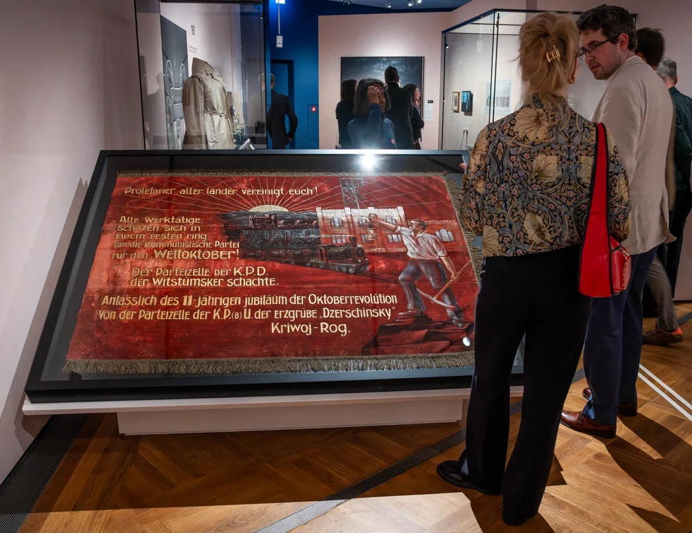 Zwei Ausstellungsgäste stehen vor einer großen, geneigten Vitrine und betrachten das darin befindliche textiles Ausstellungsobjekt, einer Art Wandteppich, auf dem rechts Eisenbahnwaggons und eine männliche Figur mit hochgekrempelten Ärmeln und einer Spitzhacke in seiner linken Hand zu sehen sind. Oben auf dem Objekt ist zu lesen "Proletarier aller Länder vereinigt euch!", darunter folgt weiterer Text.