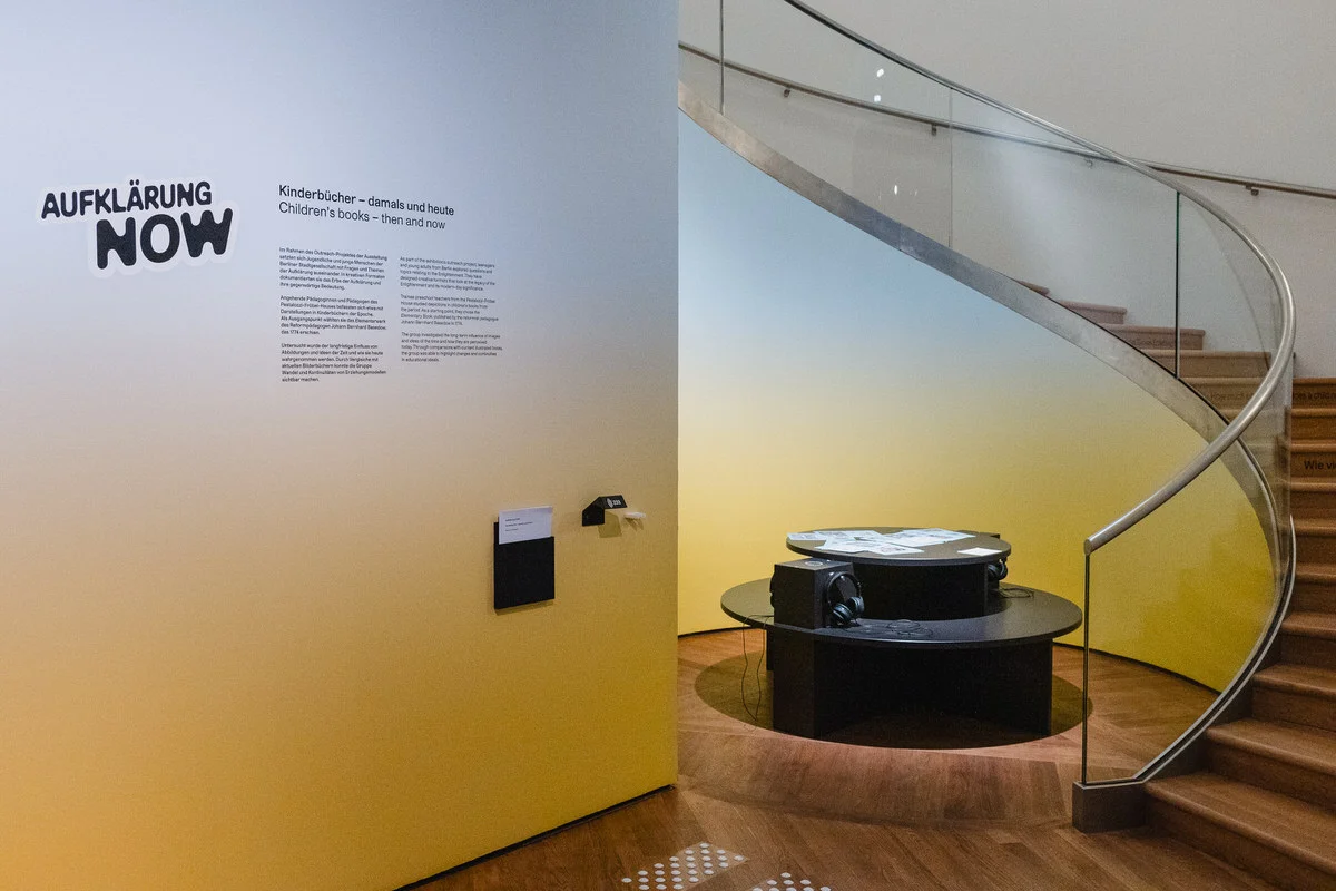 Im Vordergrund ist eine Wand mit Ausstellungstexten zu sehen. Im Hintergrund stehen auf einem runden Tisch Bücher.